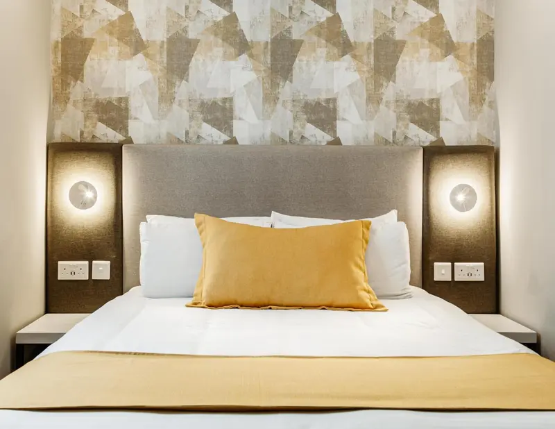 Un lit double avec des oreillers et une couverture jaune est mis en valeur par un mur texturé et des lampes de chevet.
