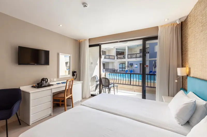 Une chambre lumineuse avec un lit blanc et un bureau donnant sur une terrasse et une piscine.