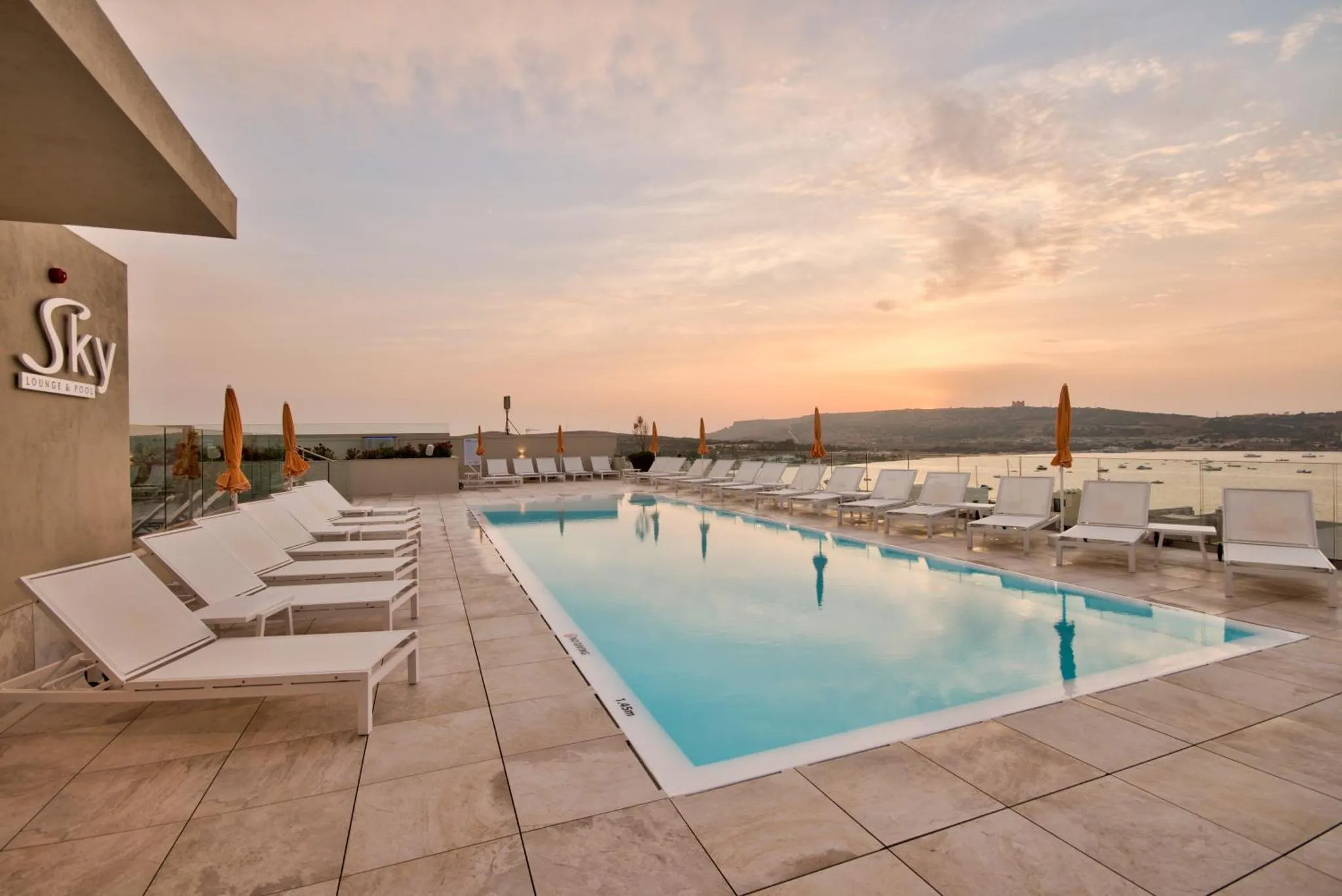Transats blancs alignés au bord d'une piscine rectangulaire avec vue sur la mer au coucher du soleil.