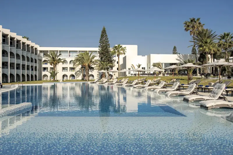 Vue d'une piscine turquoise devant un bâtiment blanc et des palmiers.