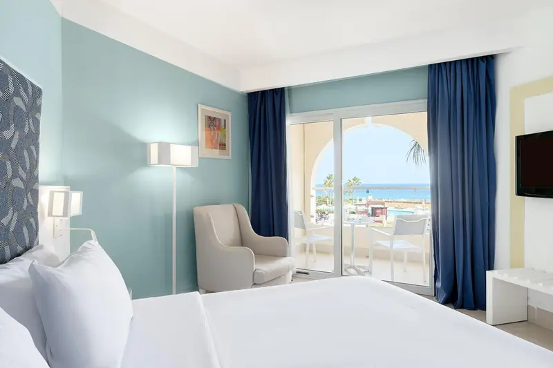 Chambre lumineuse avec un lit blanc et une vue sur la mer depuis une terrasse.