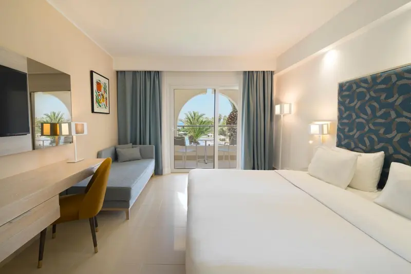 Chambre lumineuse avec un lit blanc et un coin salon près d'une baie vitrée.