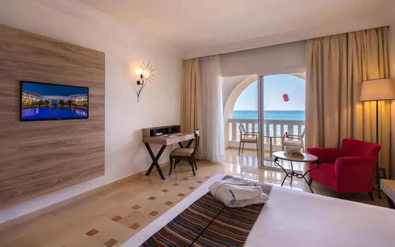 Chambre lumineuse avec bureau, fauteuil rouge et vue sur la mer.