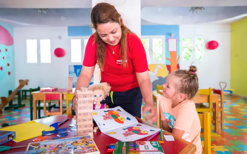 Une animatrice aide une enfant à colorier dans une salle de jeux colorée.