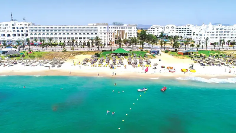 Vue aérienne d'une plage de sable fin bordée de transats et de parasols, avec une mer turquoise et un hôtel en arrière-plan.