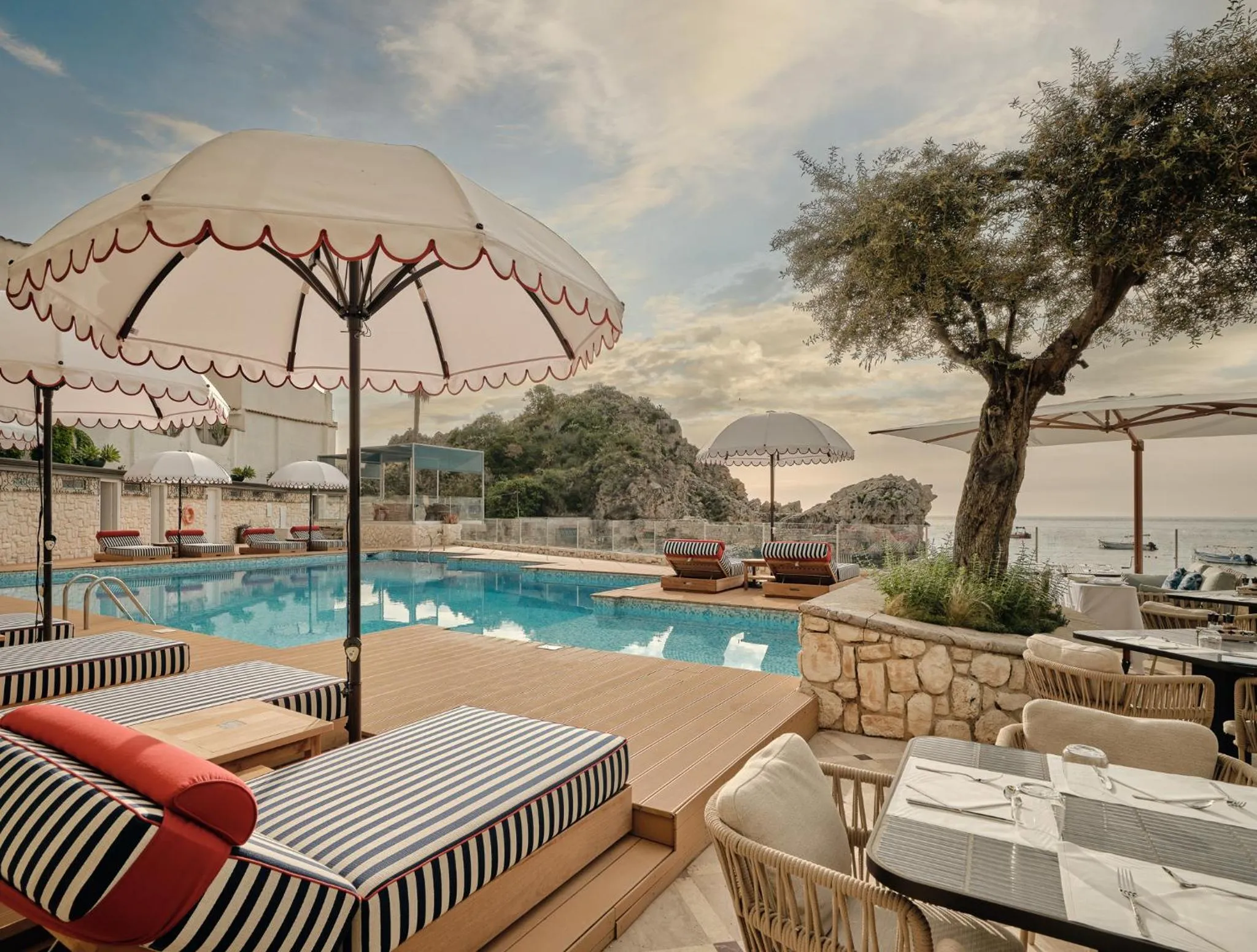 Transats rayés et parasols blancs bordent une piscine turquoise avec vue sur la mer.