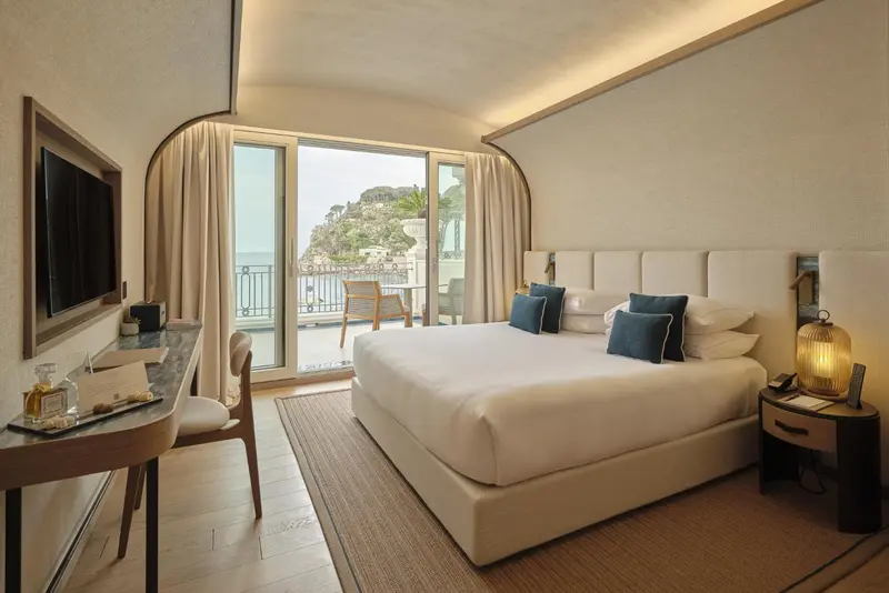 Chambre lumineuse avec un grand lit et une vue sur la mer depuis un balcon.