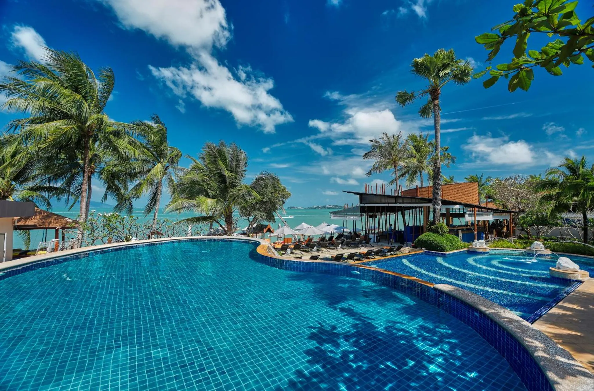 Une piscine bleue bordée de transats et de palmiers sous un ciel nuageux.