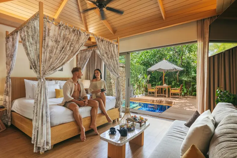 Un couple est assis sur un lit dans une chambre ouverte sur une terrasse avec piscine.