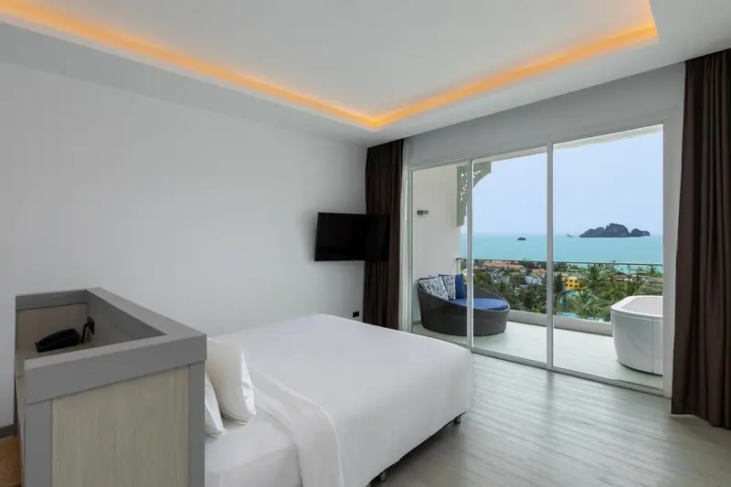 Chambre lumineuse avec un grand lit et une vue sur la mer depuis un balcon.