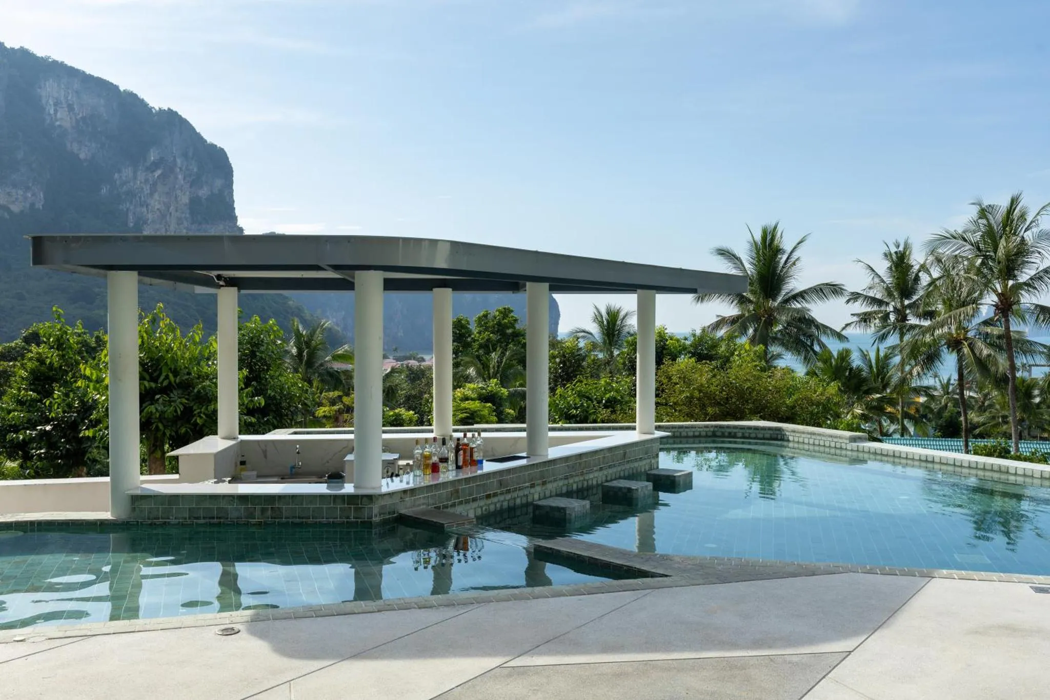 Un bar extérieur surplombe une piscine turquoise avec une vue sur la mer et la végétation.