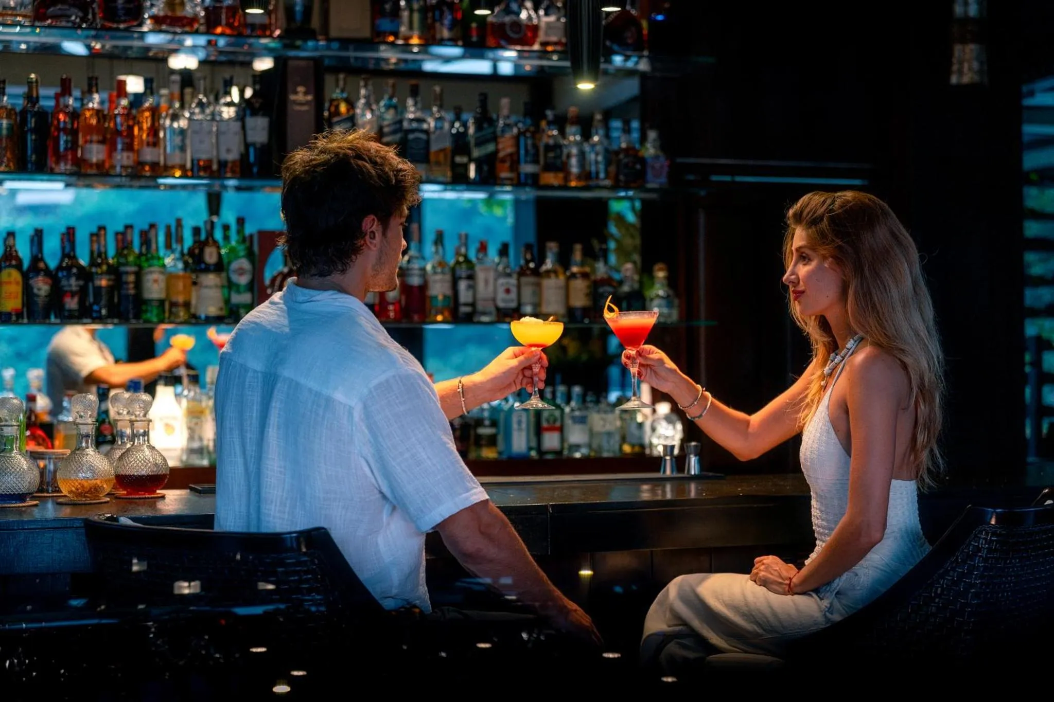 Un couple trinque avec des cocktails devant un comptoir de bar illuminé.