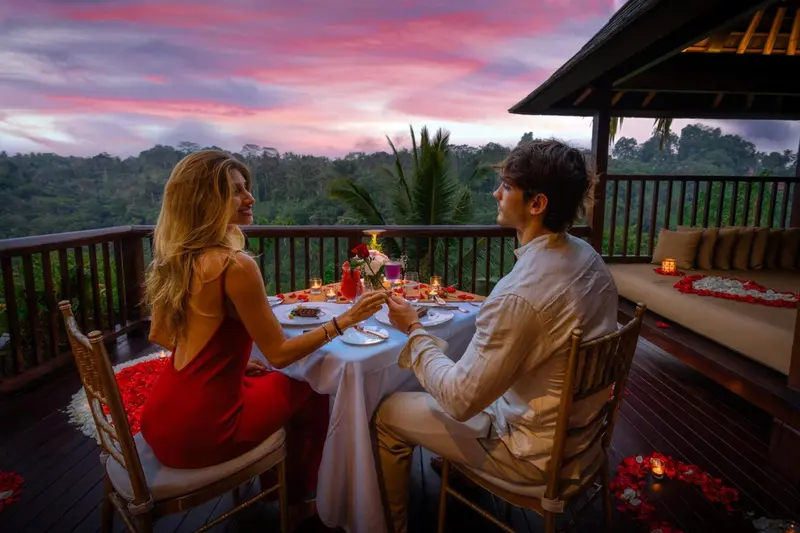 Un couple dîne à une table décorée sur une terrasse avec vue sur un paysage vallonné au coucher du soleil.