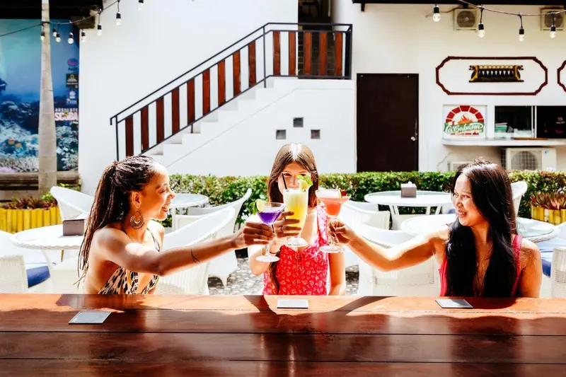 Trois femmes trinquent avec des cocktails colorés à une table en extérieur.
