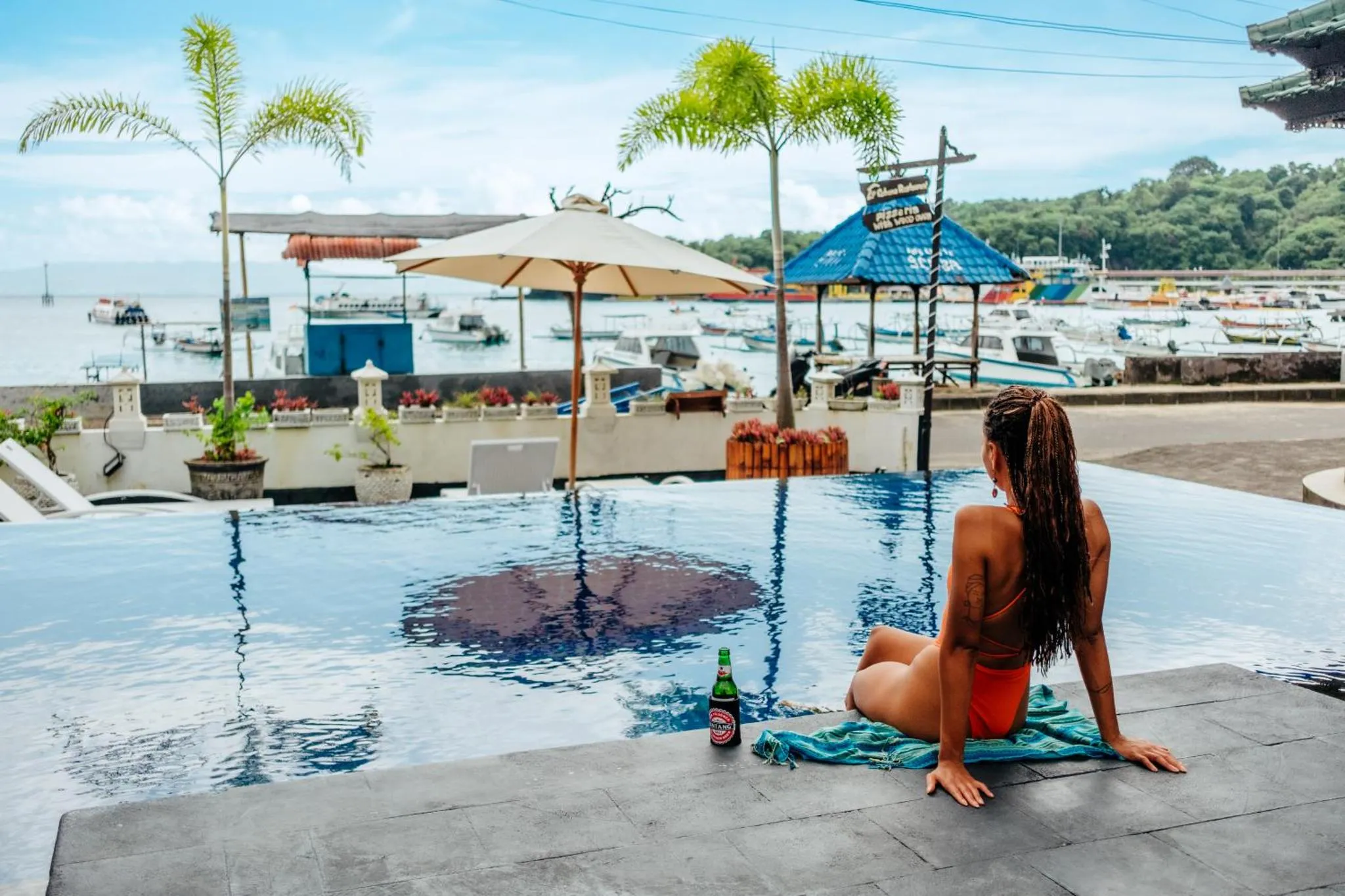 Une femme est assise au bord d'une piscine avec vue sur la mer et des bateaux.