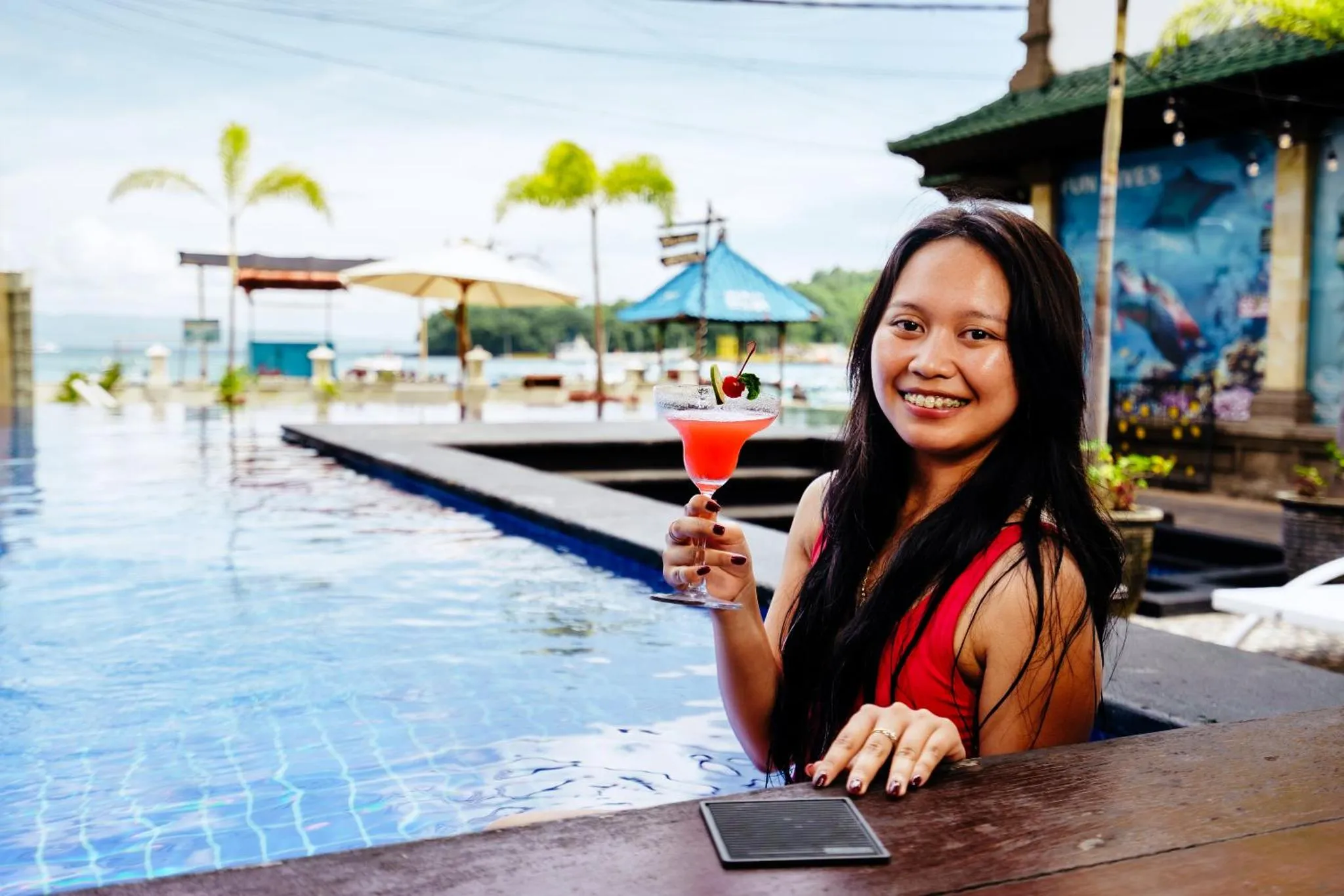Une femme souriante tient un cocktail rose devant une piscine.