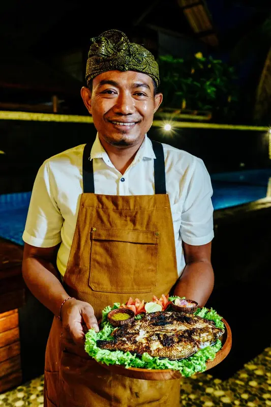 Un homme souriant présente un plat de poisson grillé sur un plateau.