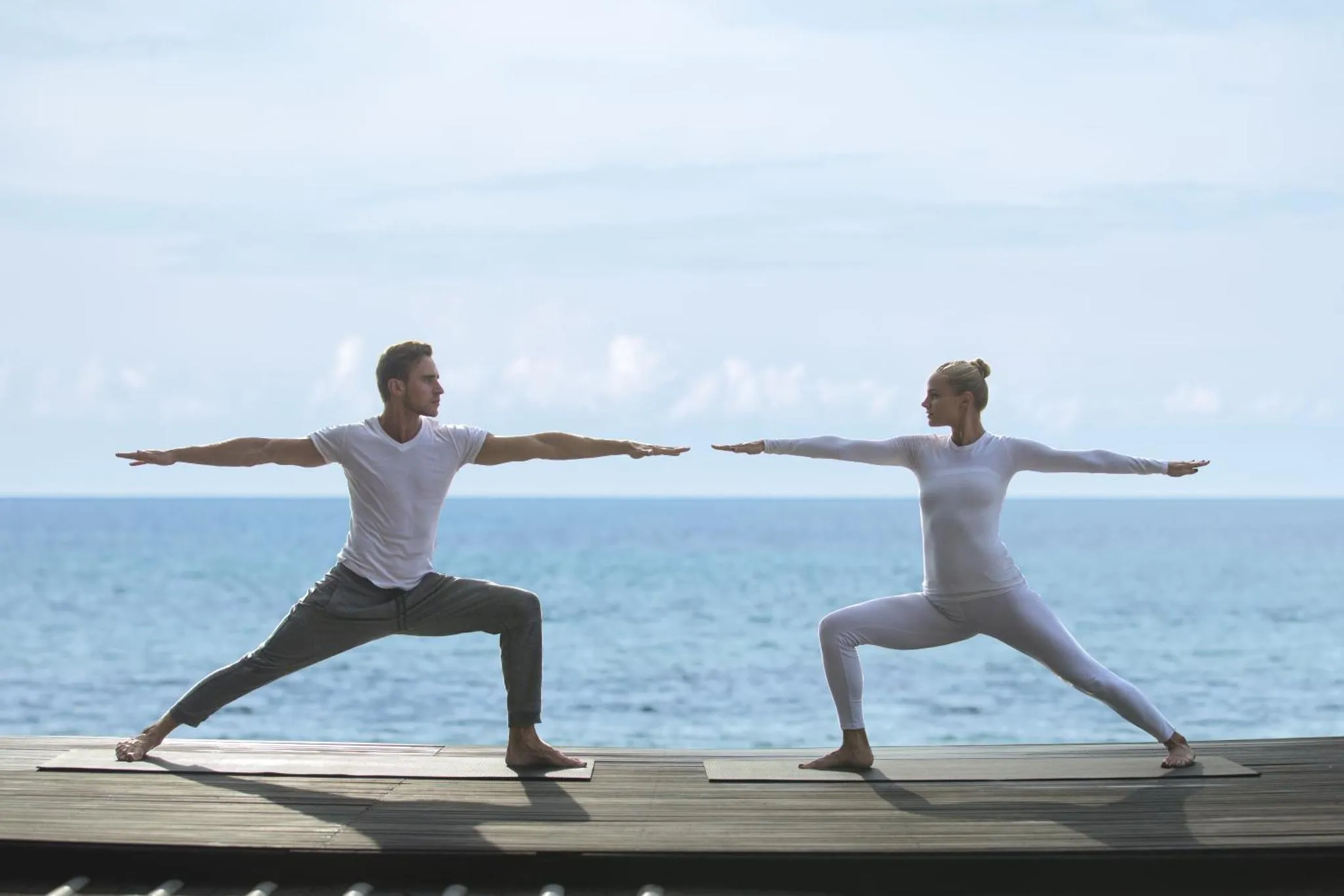 Deux personnes pratiquent le yoga sur une terrasse en bois face à la mer.