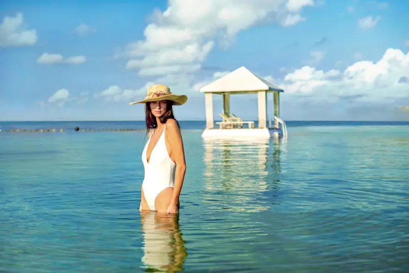 Une femme souriante se tient dans l'eau turquoise face à un ponton.
