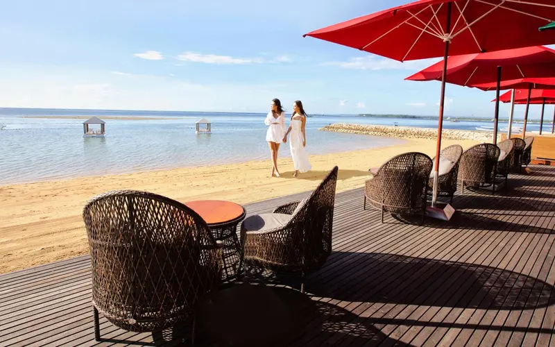 Deux femmes se promènent sur une plage de sable fin bordée de parasols rouges et de mobilier de jardin.