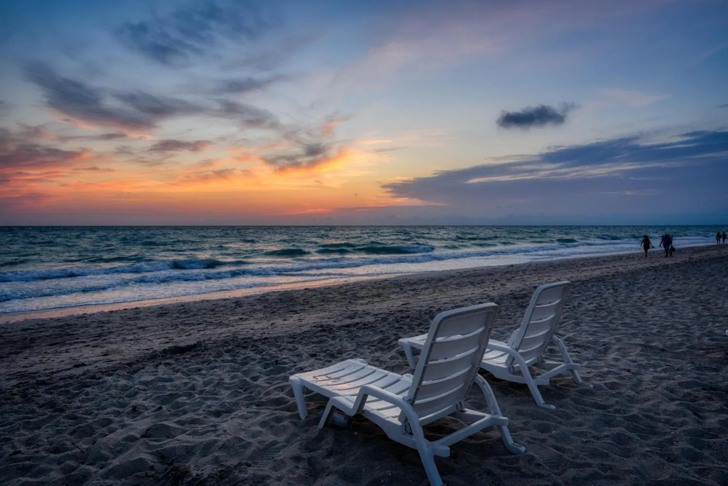 Deux transats blancs sont posés sur une plage de sable sombre au coucher du soleil.