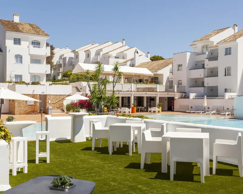 Terrasse blanche avec tables et chaises devant une piscine et des bâtiments blancs.