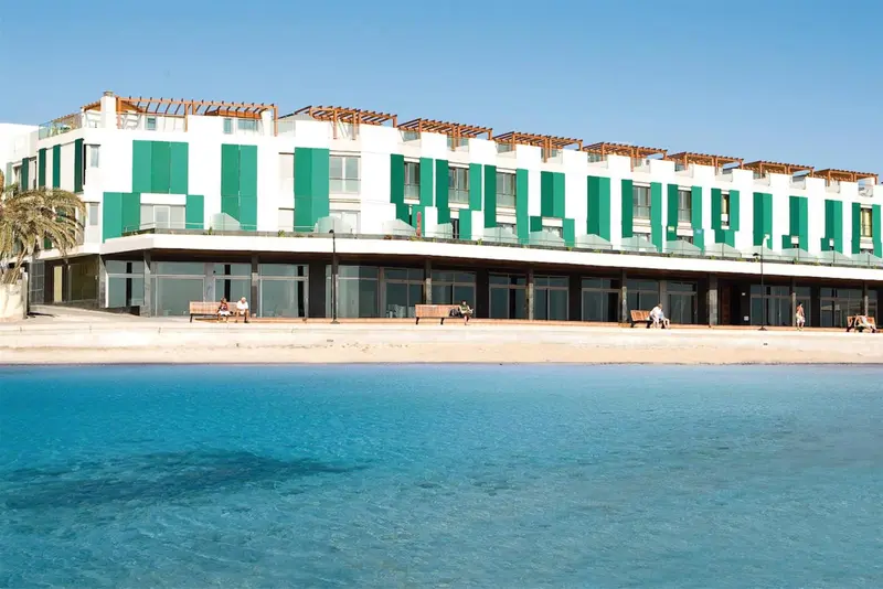 Bâtiment blanc à plusieurs niveaux avec des balcons et des persiennes vertes, au bord de l'eau turquoise.