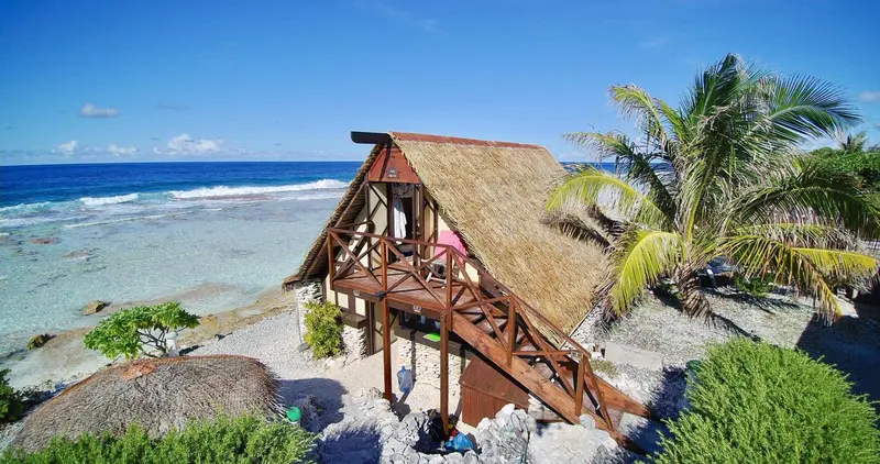 Une cabane sur pilotis avec toit de chaume surplombe une plage de sable clair.