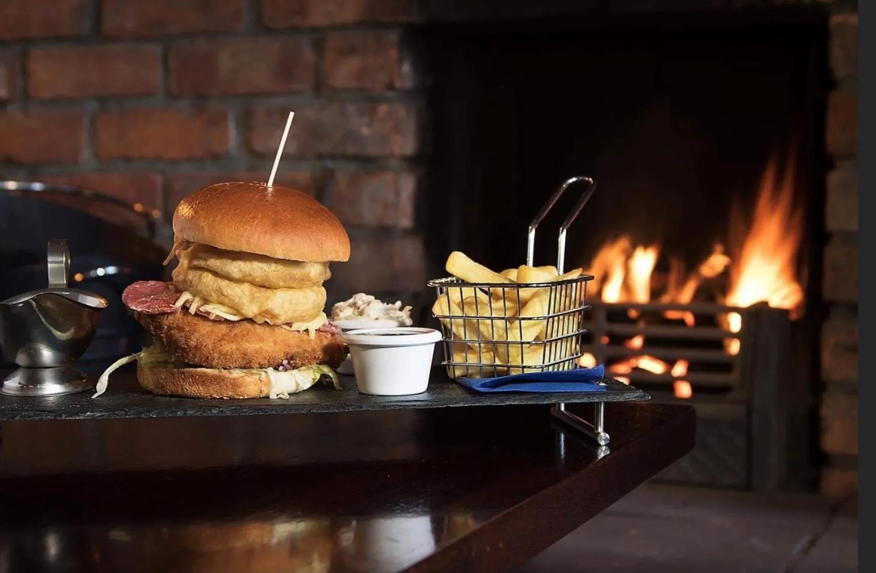 Un copieux burger et des frites sont servis sur une planche en ardoise devant une cheminée allumée.