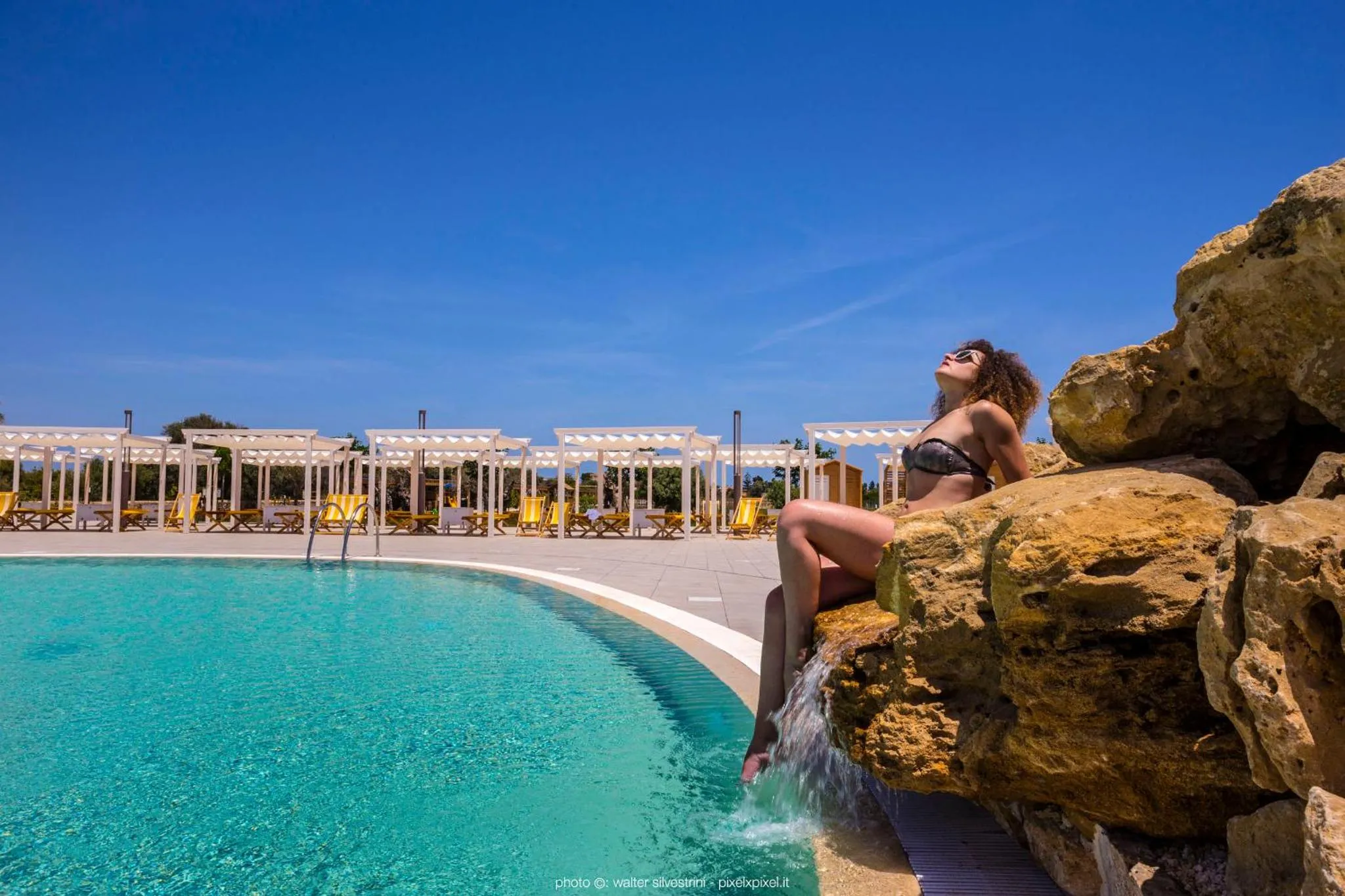 Une femme se détend au bord d'une piscine turquoise avec une cascade et des transats.