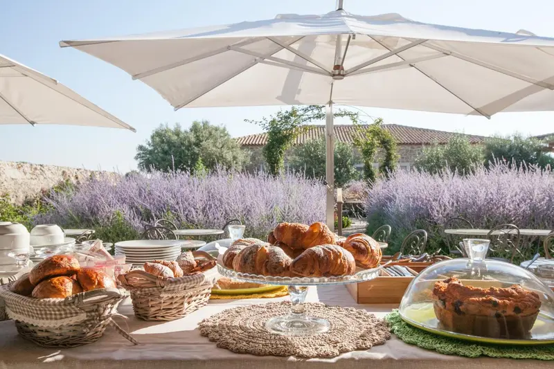 Une table garnie de viennoiseries et de pâtisseries est présentée en extérieur sous des parasols.