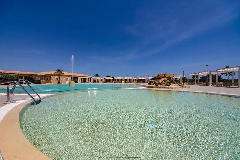 Piscine turquoise bordée d'une terrasse ensoleillée avec un bâtiment bas en arrière-plan.