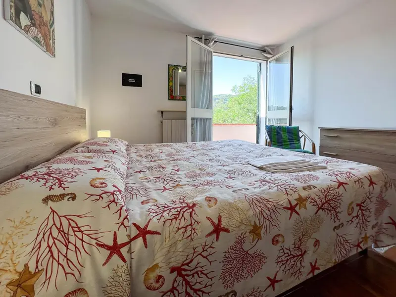 Une chambre lumineuse avec un grand lit recouvert d'une couette à motifs marins et une terrasse accessible par des portes-fenêtres.