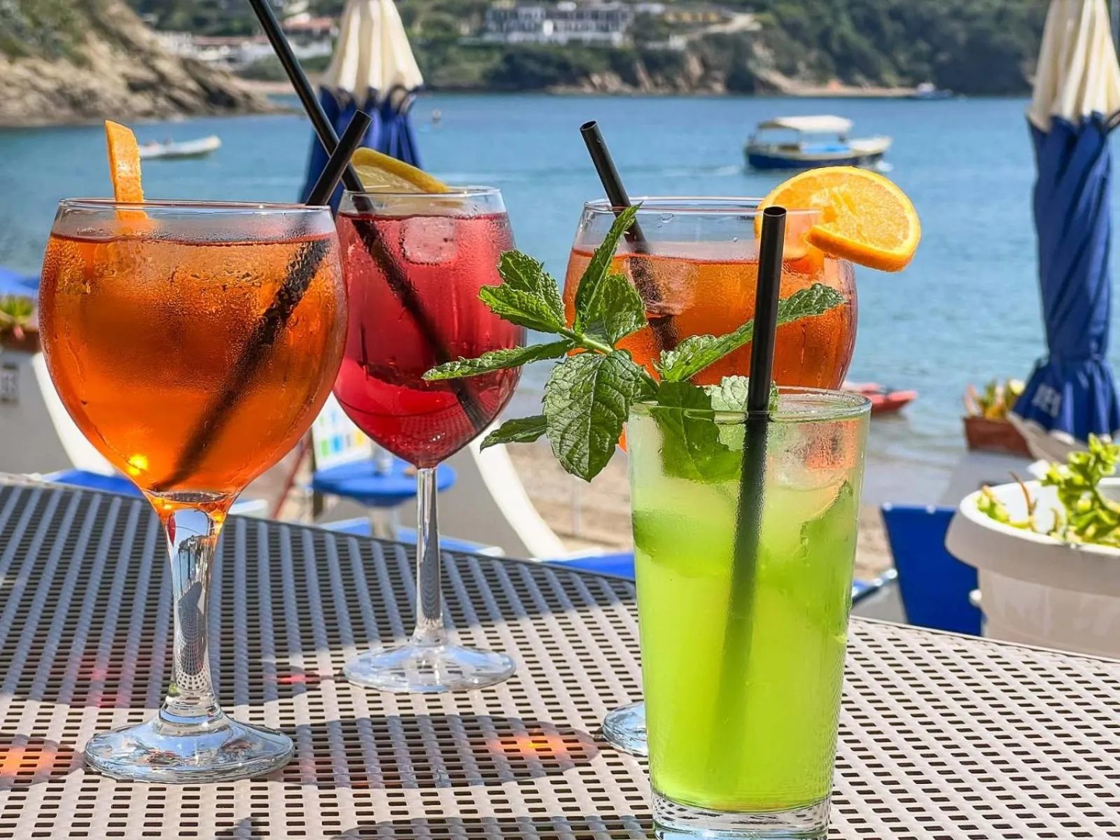 Trois cocktails colorés sont servis sur une terrasse ensoleillée avec vue sur la mer.