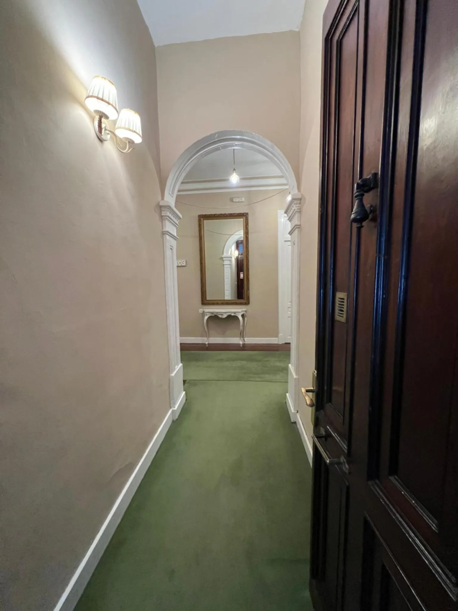 Un couloir intérieur mène à un miroir et à une porte en bois sombre.