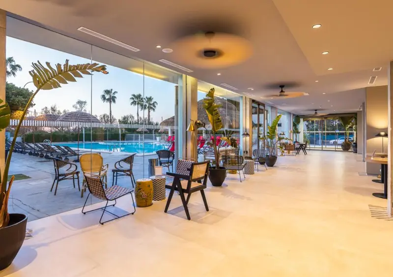 Espace de détente intérieur lumineux avec mobilier design et vue sur une piscine et des palmiers.