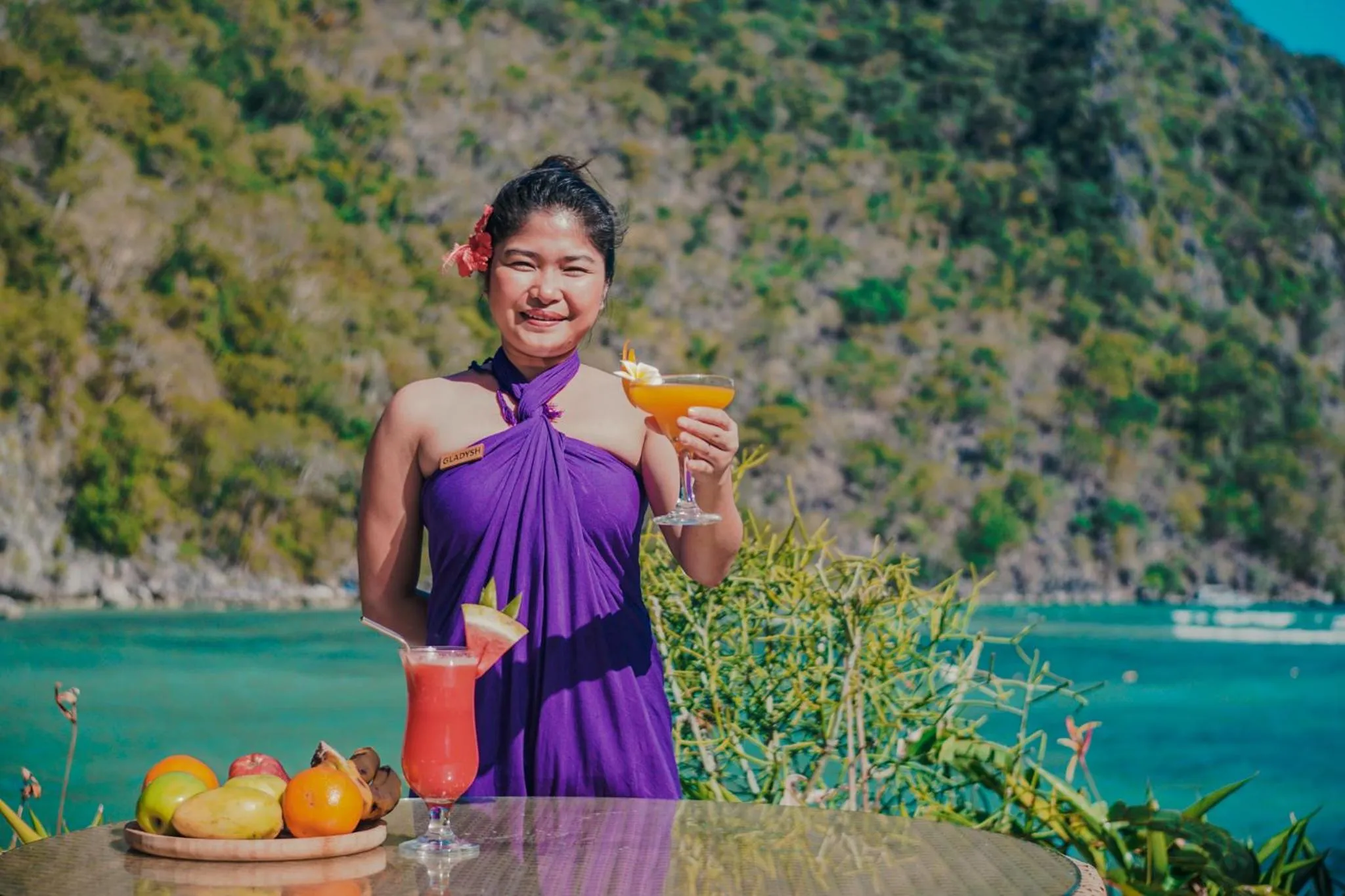 Une personne souriante tient deux cocktails colorés devant une plage tropicale.