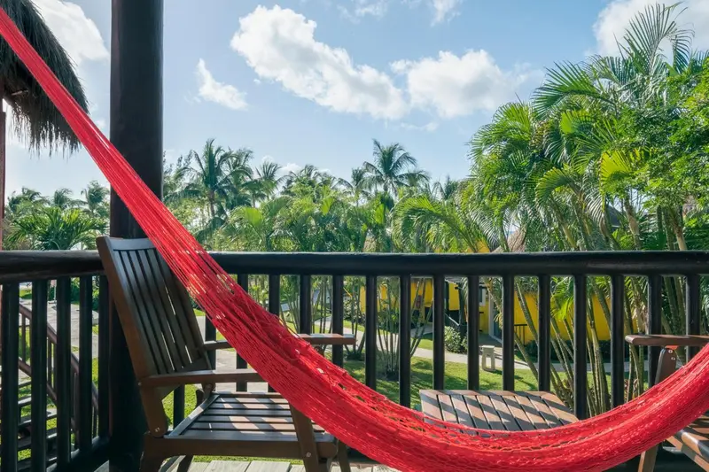 Une terrasse en bois avec un hamac rouge et des fauteuils en osier offre une vue sur une végétation luxuriante.