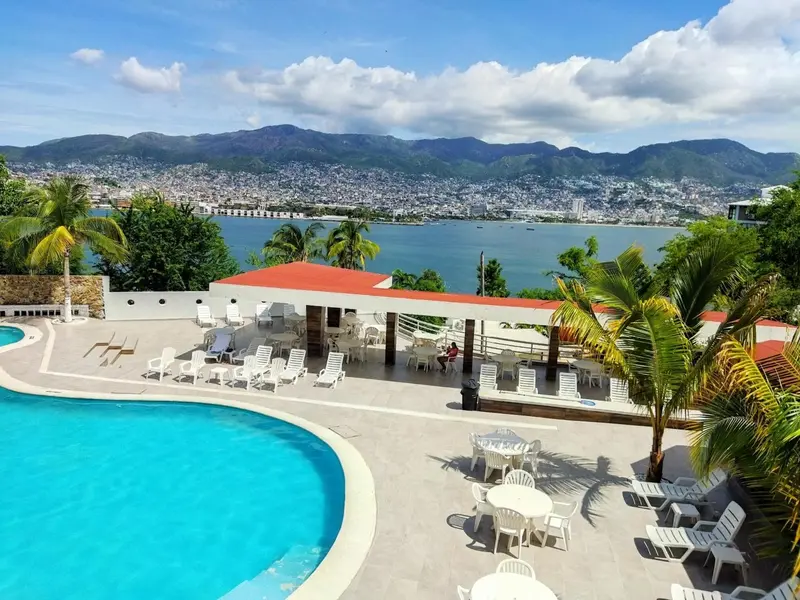 Vue aérienne d'une piscine turquoise bordée de transats et d'une terrasse ombragée avec vue sur la mer et les montagnes.