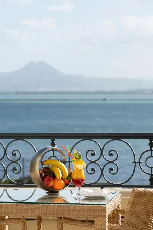 Une table avec des fruits et une boisson est posée sur un balcon avec vue sur la mer et une montagne.