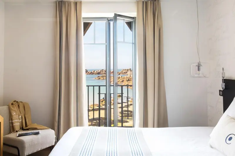 Une chambre lumineuse avec un lit et un fauteuil donnant sur une vue mer.