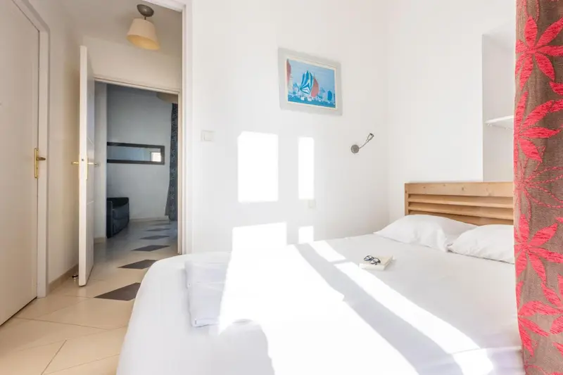 Une chambre lumineuse avec un lit fait et un canapé visible au fond.