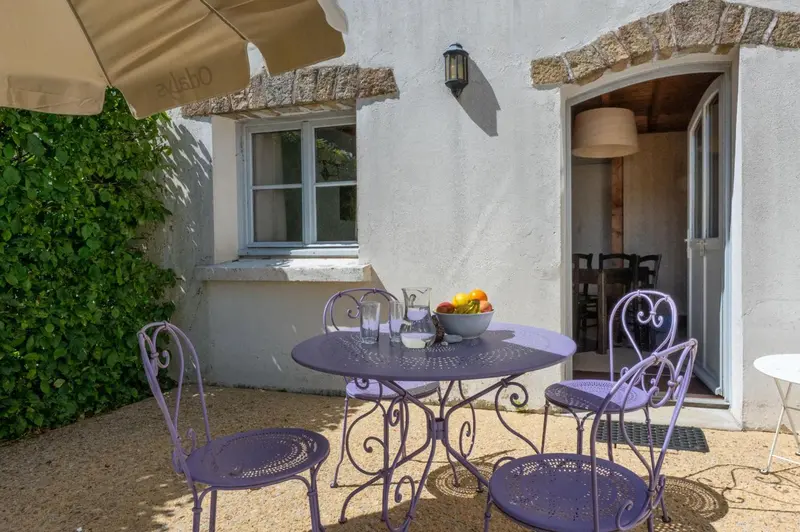 Une table et des chaises violettes sont disposées sur une terrasse en gravier devant une façade blanche.