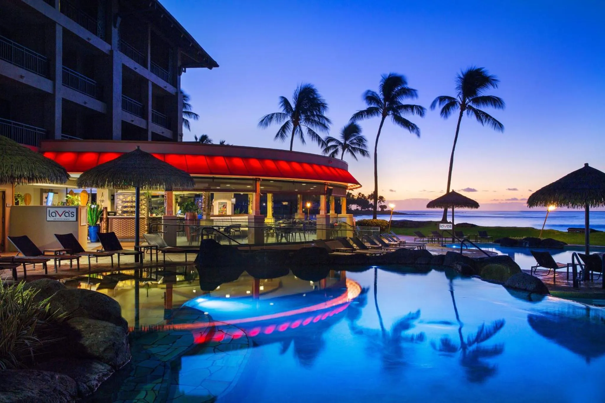Une piscine illuminée avec des chaises longues et un bar en bord de mer au crépuscule.