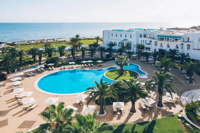 Vue aérienne d'un complexe hôtelier avec piscine et palmiers face à la mer.
