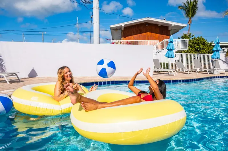 Deux femmes sourient et jouent avec un ballon dans une piscine ensoleillée.