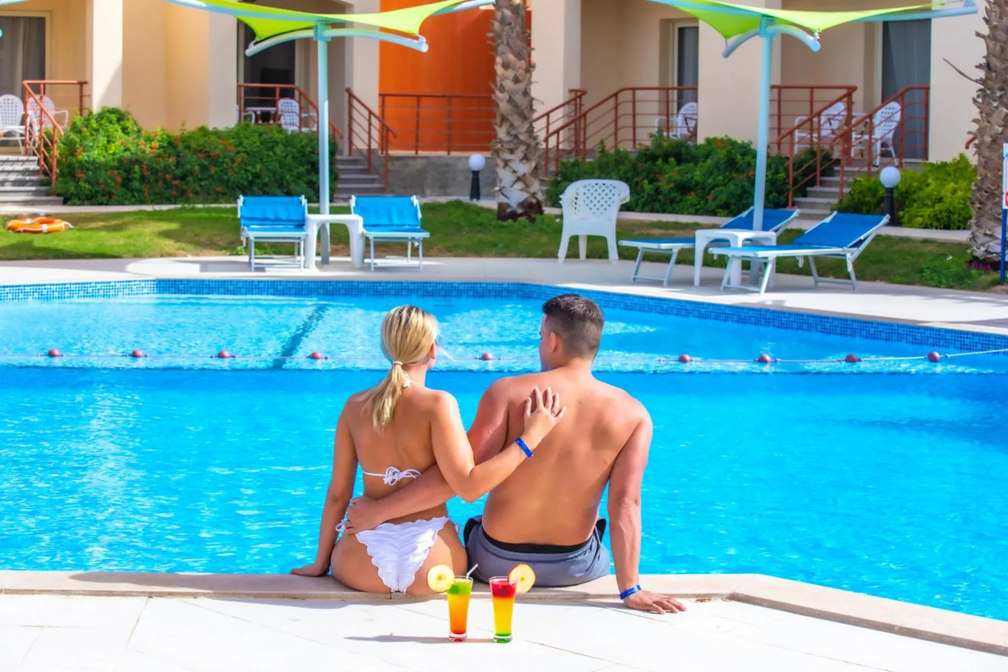 Un couple est assis au bord d'une piscine ensoleillée, avec des transats et des parasols en arrière-plan.