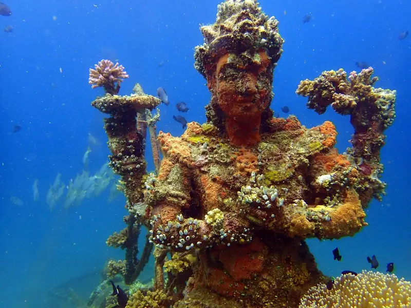 Statue coloniale recouverte de coraux et de poissons dans une eau turquoise.