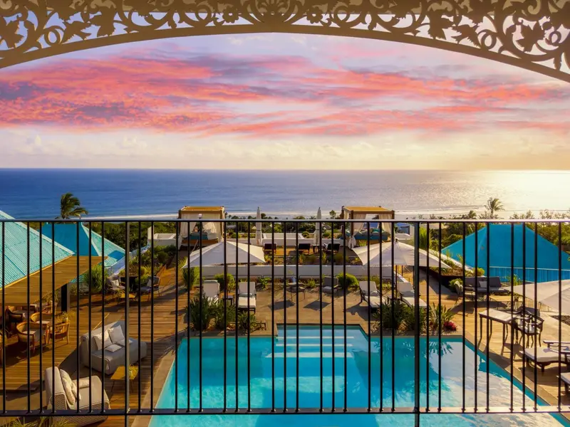Vue d'un ciel rose et orangé au coucher du soleil sur l'océan, depuis un balcon avec une piscine en contrebas.