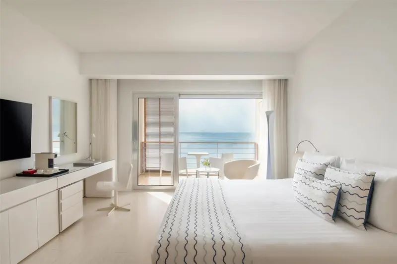 Chambre lumineuse avec un grand lit et une vue sur la mer depuis un balcon.